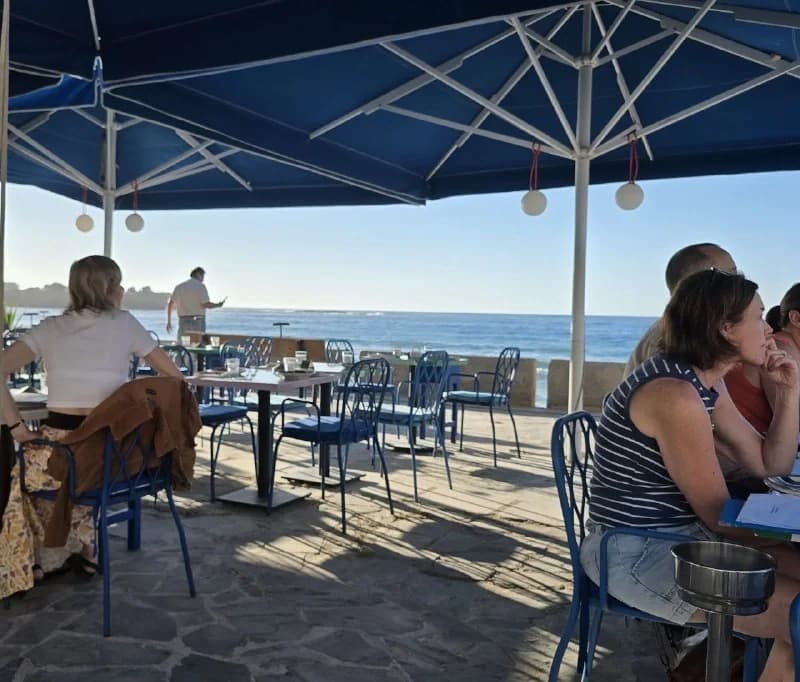Travelers gathered beneath umbrellas at a waterside café